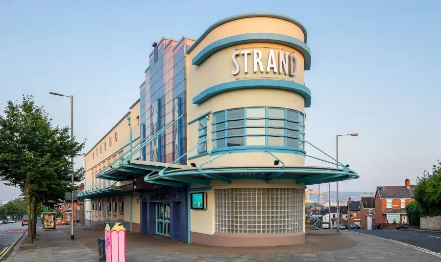 An art deco building located on the apex of two roads. The word Strand is in silver lettering on the top portion of the curved exterior.