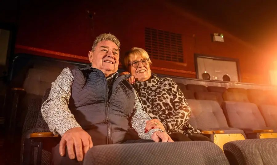 Two people are seated on a blue double cinema seat in Strand Arts Centre screen one. They are facing towards the screen and smiling.
