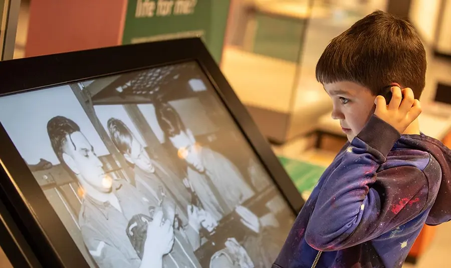 A child looks at a screen with a black and white image, holding a speaker up to their ear