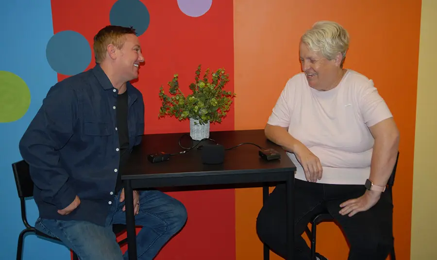 Two people laughing with a recording device on the table in front of them