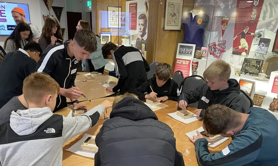 Two tables of teenagers focus on a crafting task. Historic kits and footballing ephemera are displayed in glass cabinets behind them.