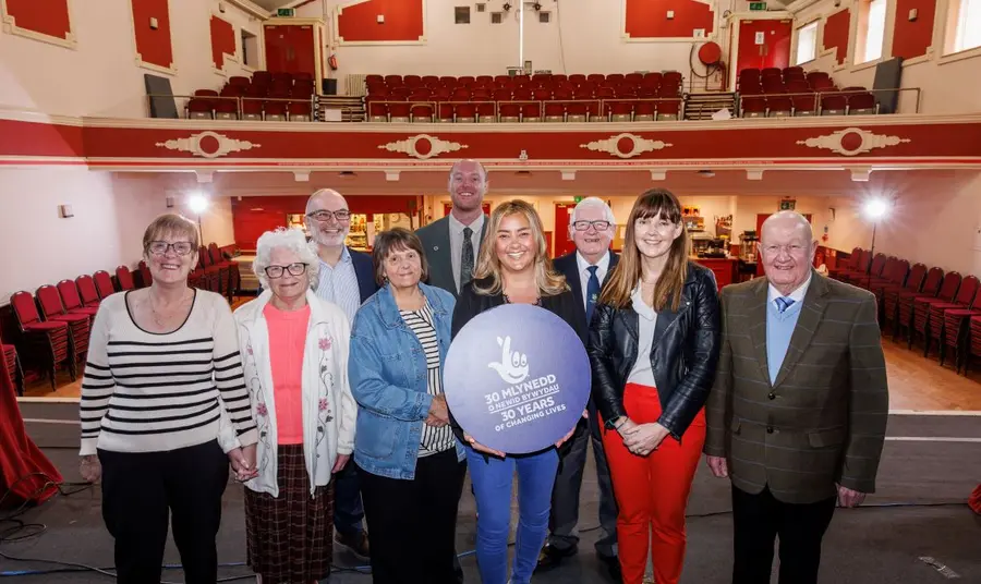 A group of local people stand on the stage of the Welfare Hall, with the auditorium behind them. In the middle, a young woman holds the National Lottery Heritage Fund blue roundel.