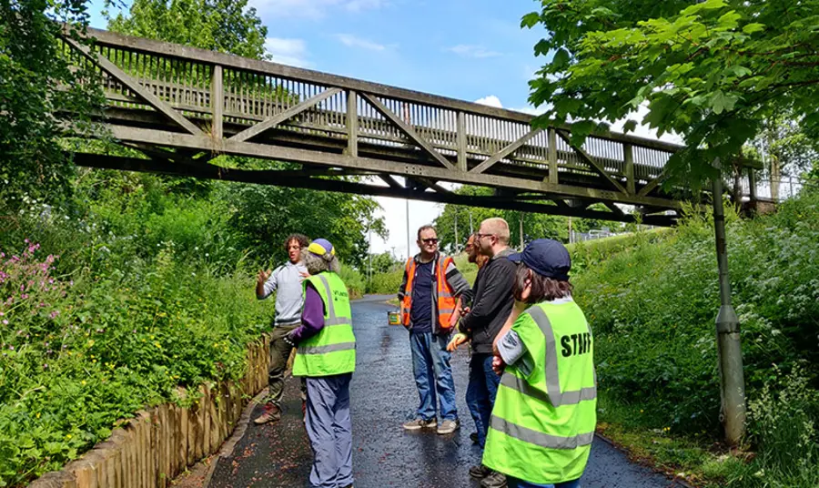 A group of people standing in a lane above them is a wooden bridge, the lane is enclosed by greenery covered embankments.