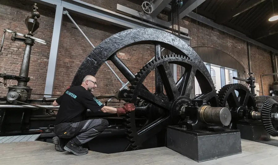 A volunteer crouches to attend to the mechanics of enormous overlapping cog wheels, part of a standing engine.