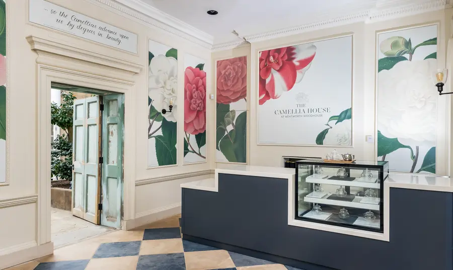 A cafe counter inside a room with a chequered tiled floor. Panels are on the wall behind the counter with illustrations of camellias.