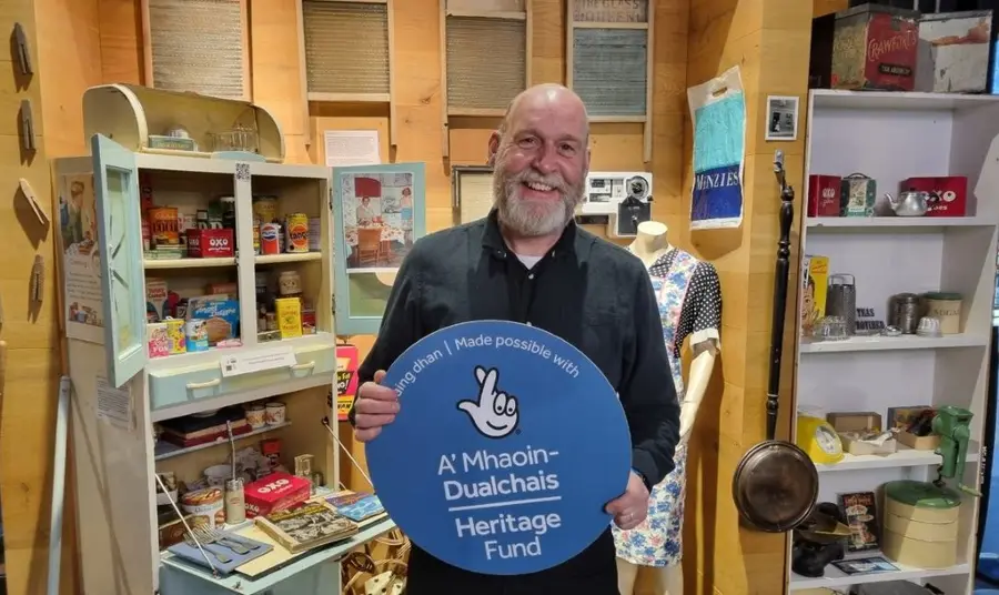 Russell Clegg holds a Heritage Fund roundel while standing in front of a display of nostalgic items connected to cooking and kitchens.