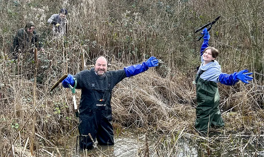Two people wearing waders wave from an overgrown pond.
