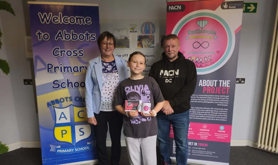 A child holding an award standing with two adults in front of banners with the Rathcoole Memories and Abbots Cross Primary School logos