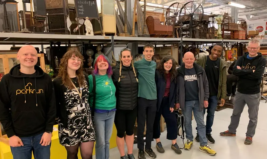 A group of men and women of various ages pose for a photo in a collections store full of antique furniture and artefacts.