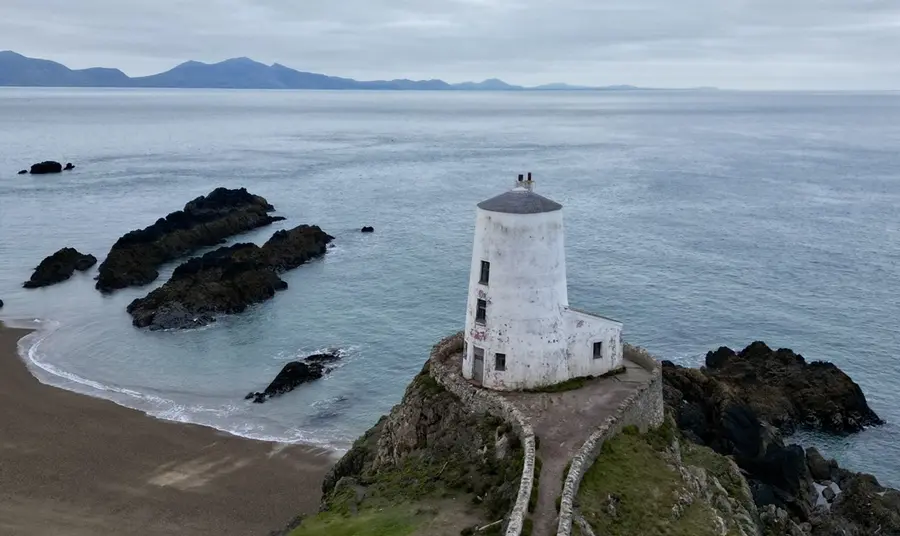 A lone white lighthouse sits atop a stack of rock on the craggy shoreline of Ynys Mon, looking out over the Menai Strait.