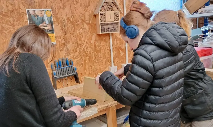 On a workshop bench, one person operates a drill as another - wearing headphones, holds the structure of a wooden bird box steady for drilling.