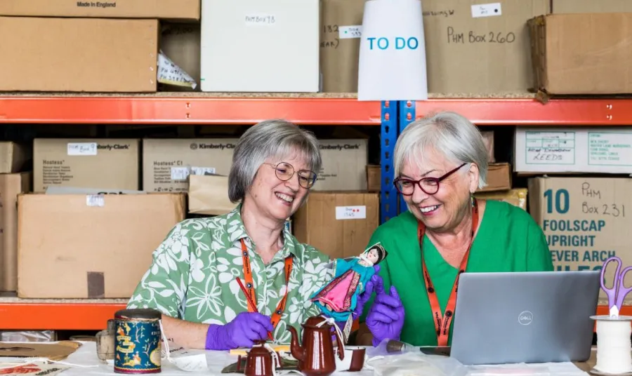 Barbara and Fiona, two smiling women both with white hair and glasses, sit at a desk in the open store and examine pottery and homewares from the collection.