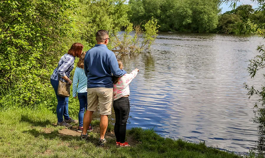 A family of two adults and two children standing on the banks of a river. One of the children is pointing at the water.