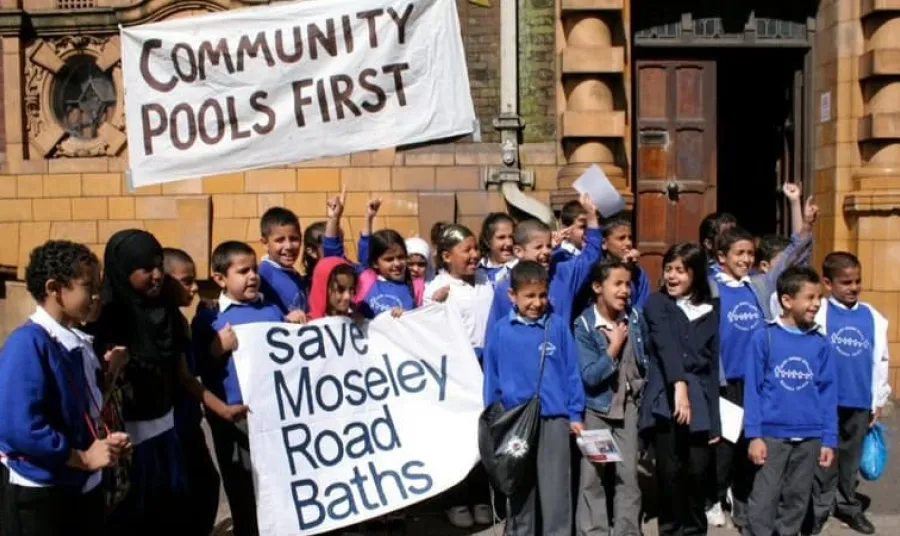 School children hold a banner reading 'save Moseley Road Baths' in front of a historic stone building