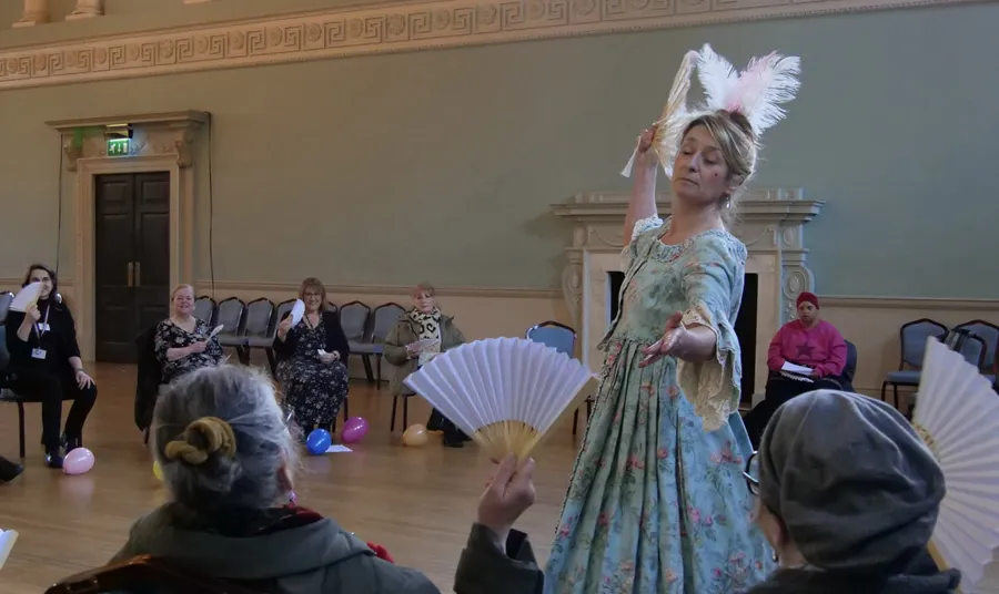 A person wearing an 18th-century style green floral gown and feathered headpiece performs a dance routine to an audience inside a historic hall