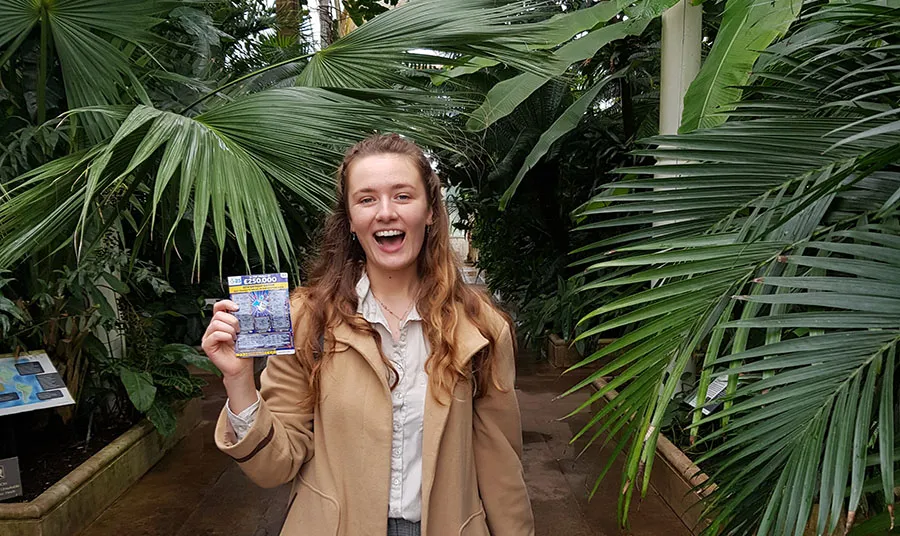 A person visits Kew Gardens Palm House and holds a National Lottery scratchcard at the camera.