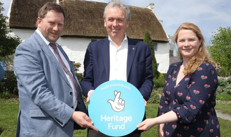 Barry Nielson, Paul Mullan and Isabel McKernon stand together in front of a thatched cottage. They hold between them a blue roundel bearing the Heritage Fund logo.