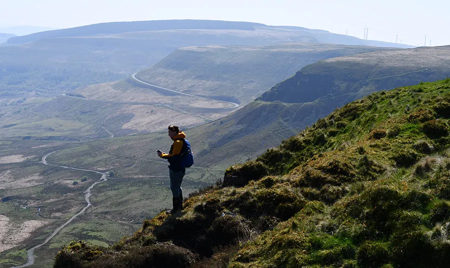 Mae dyn sy'n gwisgo cot felen gyda backpack las yn sefyll ar ochr bryn wedi'i amgylchynu gan fryniau tonnog Cymreig