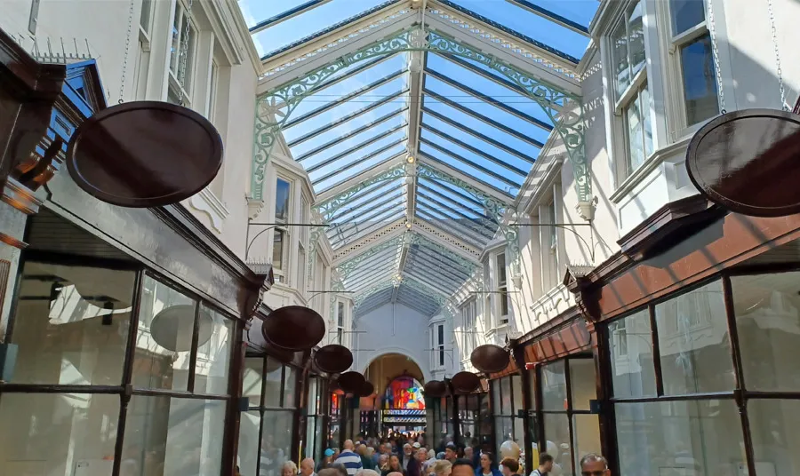 The central walkway of Dewsbury Arcade is full of shoppers with a bright blue sky seen through the intricate glass roof