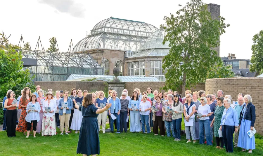 a choir sing in a garden in front of a Victorian glass and ironwork Palm House