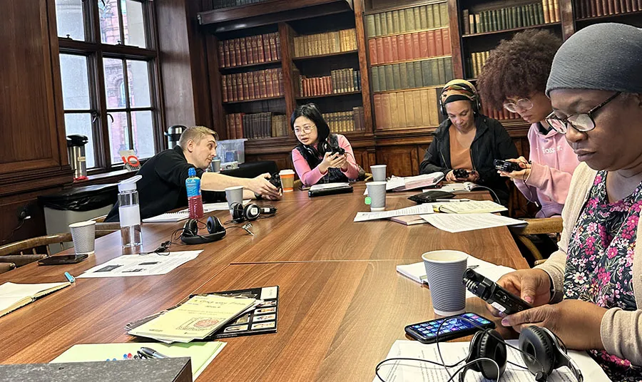 Five people sit in a library around a table covered with recording equipment and documents