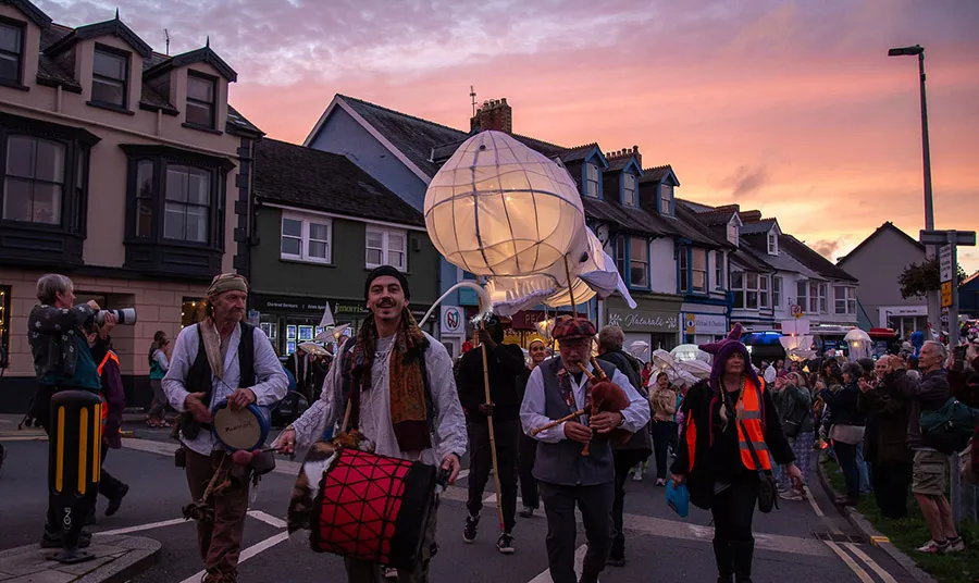 A parade at sunset featuring drummers, pipers and a whale-shaped lantern