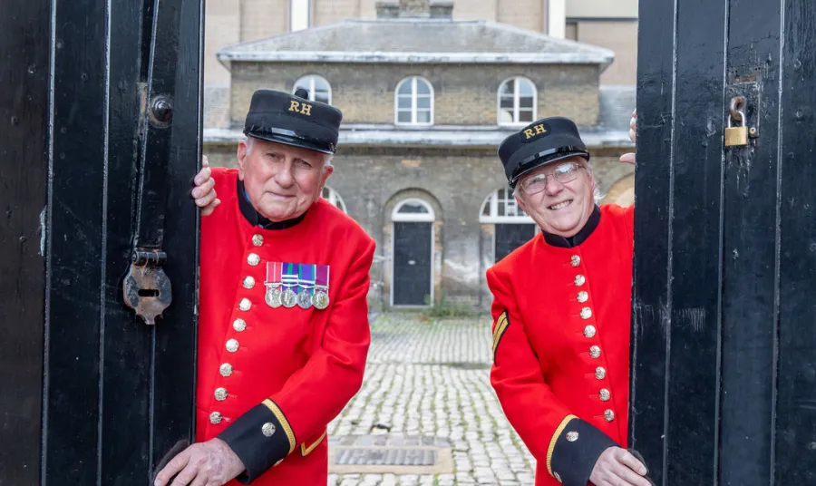 Two people wearing scarlet Chelsea Pensioners' uniform open stable doors at Royal Chelsea Hospital, London with historic building and courtyard behind