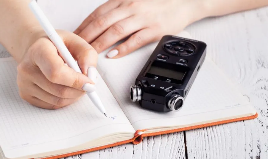 A voice recorder on a table next a a notebook with someone getting ready to take notes