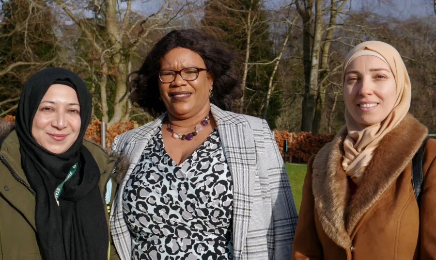 Three women from diverse ethnic communities are stood and smiling at the camera