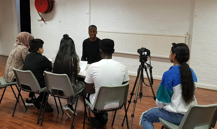 A group of young people sit a table interviewing another young person, with another person to the side operating a camerawith 
