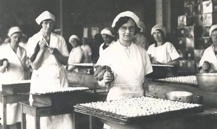 Women wearing uniforms, stood at tables in a factory, making biscuits.