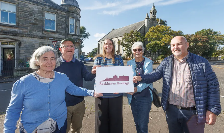 five people holding a Buckhaven Heritage sign. Behind them a stone church can be seen