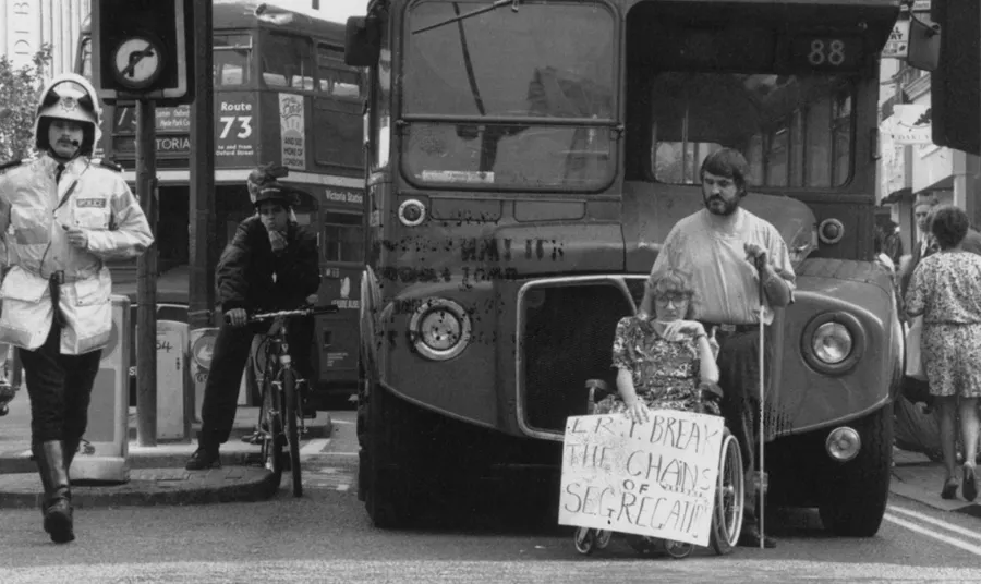An archive image showing a person in a wheelchair holding a sign in front a double double bus. There is a police officer on the left hand side.