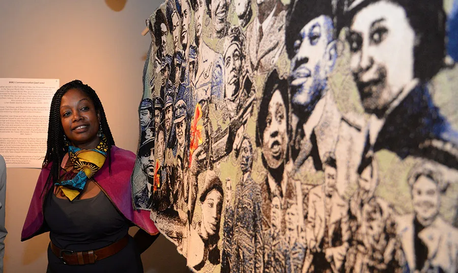 Paula Ogun Hector stands beside a woven tapestry made up of portraits of Black Britons from the Second World War