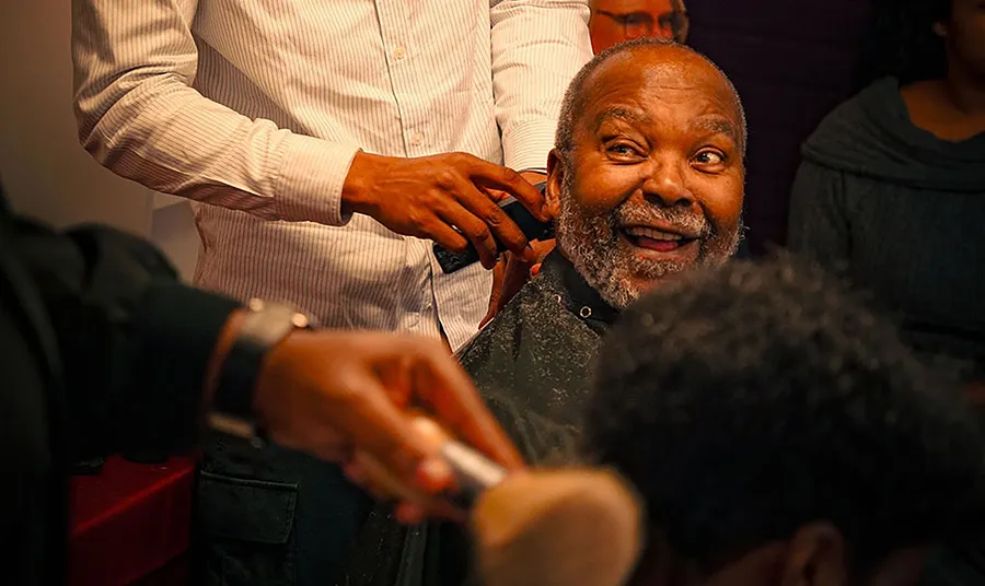 A person looks at the camera and smiles while a barber trims their hair