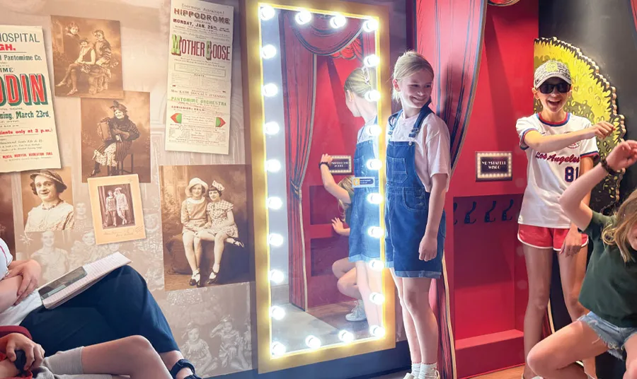 Children pose in a theatre-themed exhibit area with an illuminated dressing room mirror