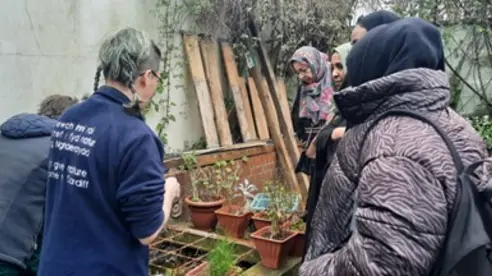 A group of women wearing headscarves listen to a wildlife educator talk, standing around a raised pond