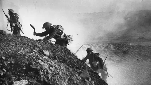 A black and white historical photo of Gurkha soldiers climbing a rocky hill. The khukuri (knife) of one of the soldiers is visible.