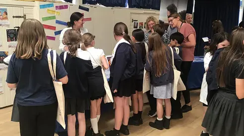 Schoolchildren at a stall about the project. A person in a white lab coat stands behind the table talking to the children.