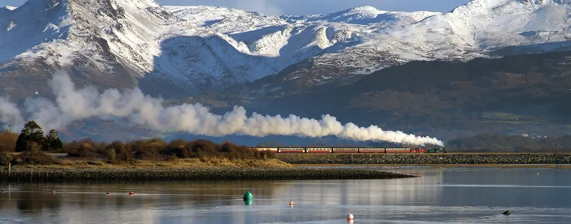 A steam train travelling next to water with snowy mountains behind