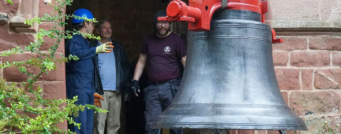 A church bell being lifted by a crane while three people look on from a doorway