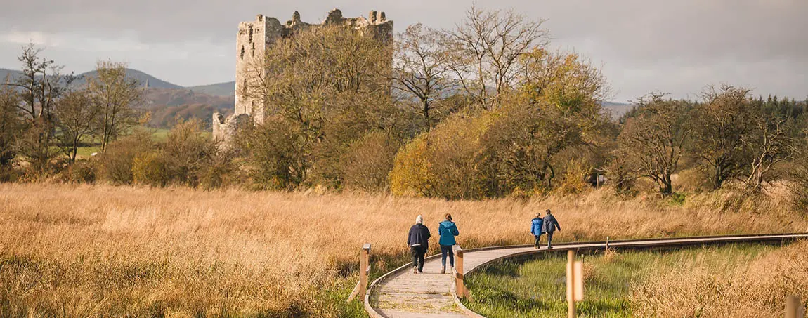 People walking on a winding wooden boardwalk through a field in front of a castle