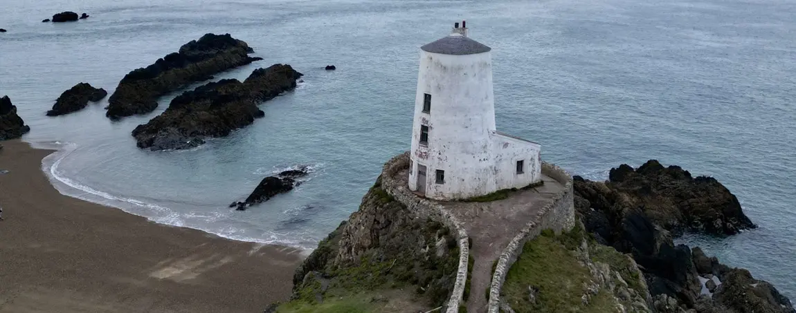 A lone white lighthouse sits atop a stack of rock on the craggy shoreline of Ynys Mon, looking out over the Menai Strait