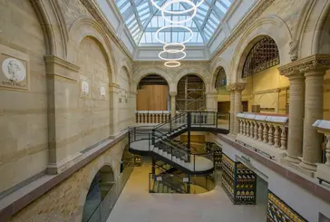 A Victorian-style interior features an exposed staircase central but at the back of the space. A balcony can be seen on the right-hand side and modern, circular lights hang from the ceiling.