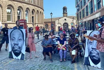 People sit outside listening to one person speaking with posters of David Oluwale around them