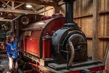 Two visitors investigate a red tank engine in a wooden shed at the Ffestiniog and Welsh Highland Railway.