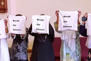 Five women stand side by side with white sheets covering their faces bearing the words "break the silence"