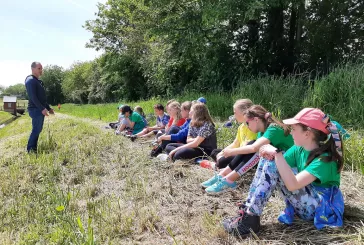 A group of children sitting down in front of a row or trees, listening to someone giving a talk