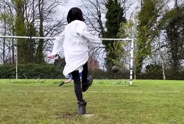 A footballer in a long white shirt and heavy work boots takes a penalty kick.