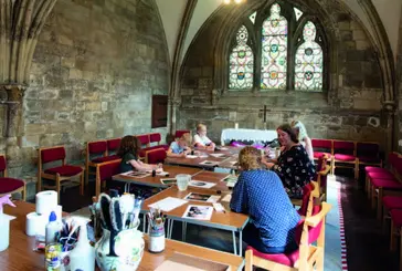 Three adults and three children are sat at tables in an Abbey hall doing crafts.